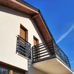 Nowoczesna, czarna balustrada balkonowa z poziomych profili na tle błękitnego nieba. Ujęcie z dołu, eksponujące geometrię i minimalistyczny design.