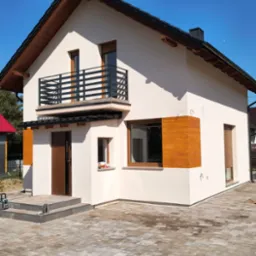 Nowoczesny dom z balkonem i czarną, metalową balustradą. Elewacja w odcieniach bieli i drewna, bruk na podjeździe. Błękitne niebo w tle.