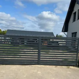 Nowoczesna, pozioma brama wjazdowa w kolorze grafitowym, z widocznym domem w tle i fragmentem ogrodzenia z siatki po lewej stronie.
