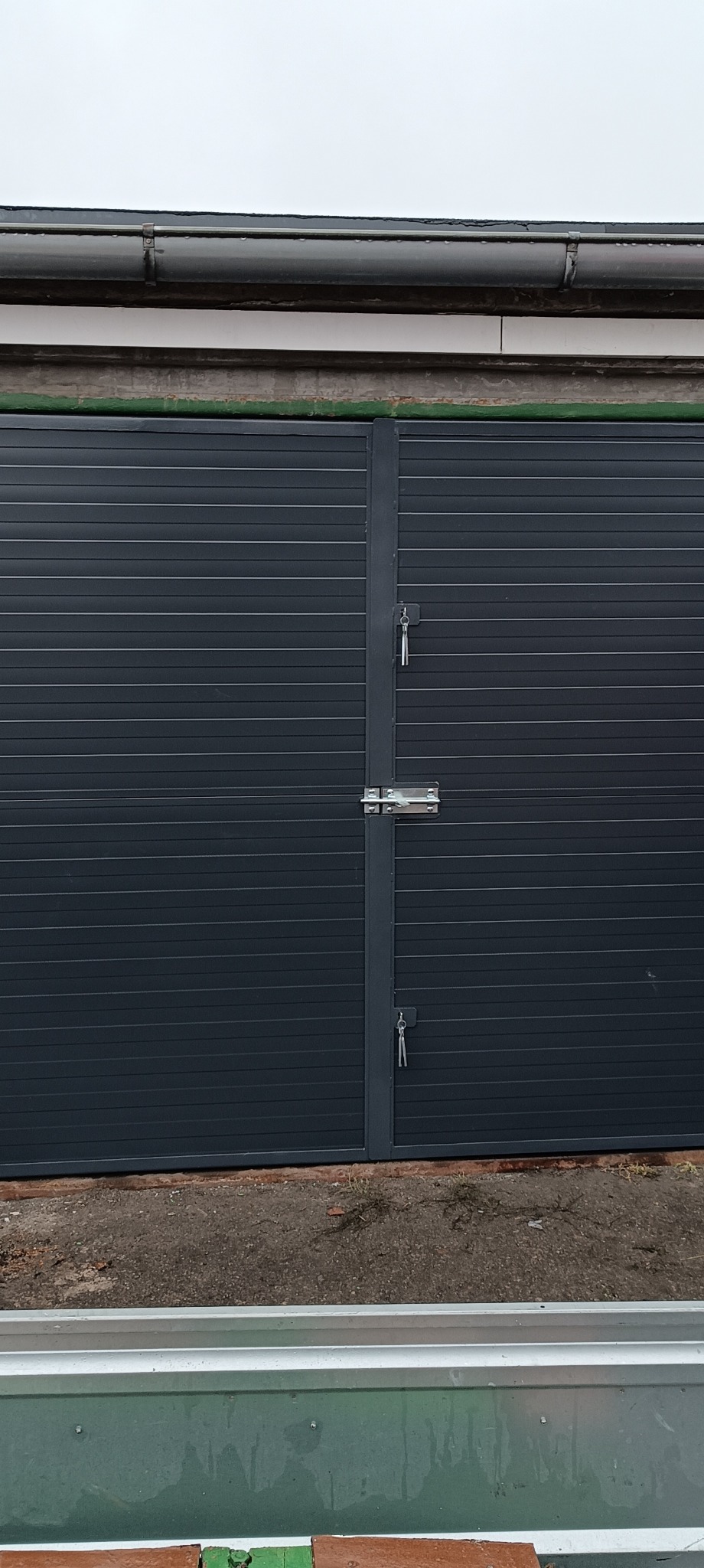 Szara, poziomo żłobkowana brama garażowa z zamkiem na klucz, widoczna z bliska, z fragmentem rynny w tle.