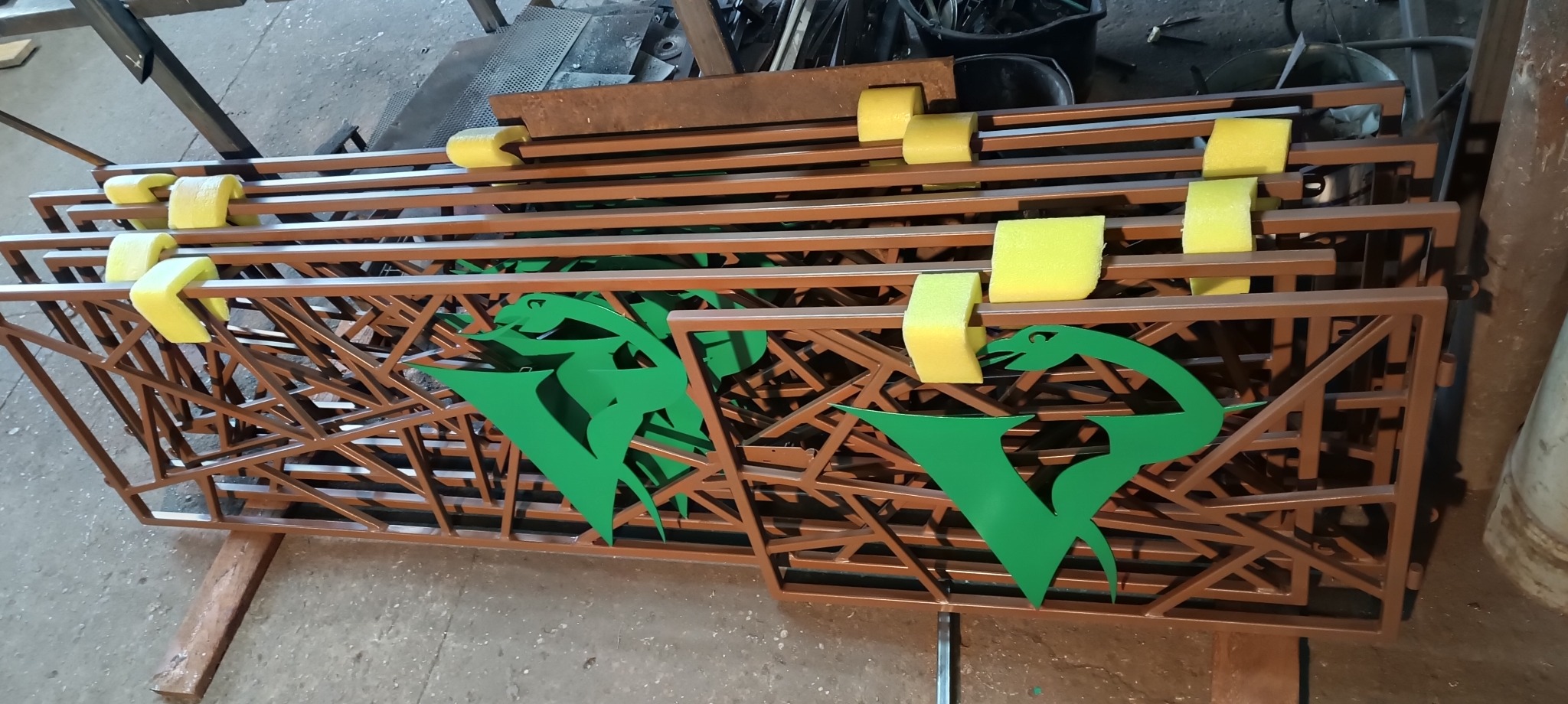 Stos brązowych, metalowych przęseł ogrodzeniowych z zielonym, wyciętym laserowo motywem roślinnym i żółtymi przekładkami ochronnymi z pianki, w warsztacie.