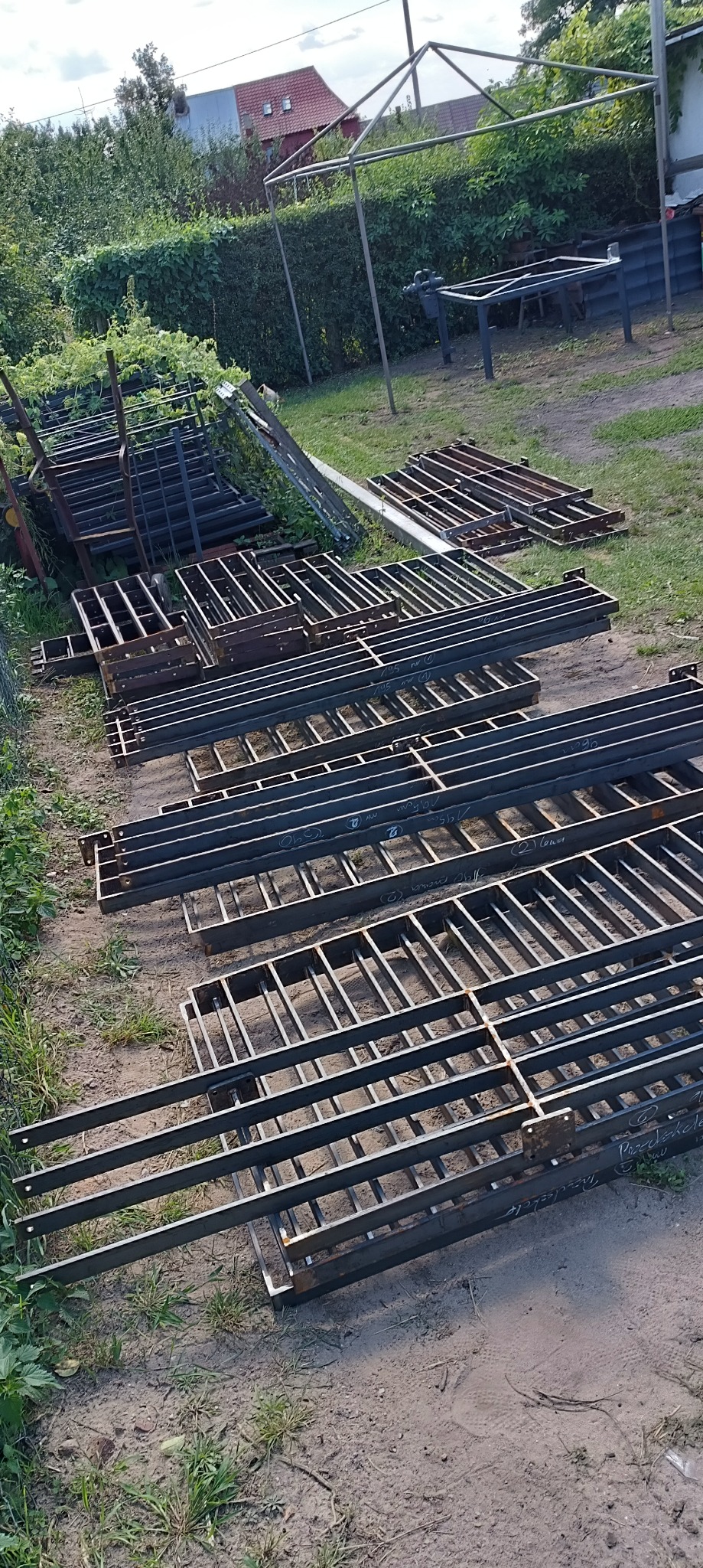 Stalowe konstrukcje kratowe, prawdopodobnie elementy bramy lub ogrodzenia, ułożone na ziemi na zewnątrz, z widocznymi spawami i oznaczeniami, w tle zieleń i częściowo widoczna zabudowa.