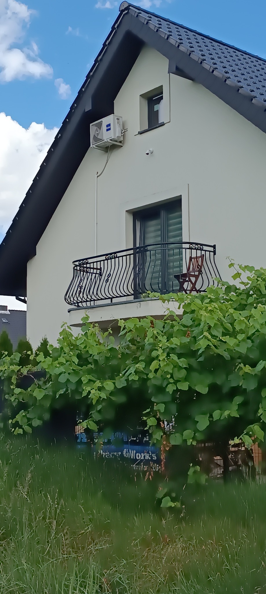 Biały dom z czarnym dachem i balkonem z dekoracyjną, kutą balustradą, częściowo zasłoniętą przez bujną zieleń winorośli. Na balkonie stoi krzesło. Na elewacji widoczny klimatyzator.