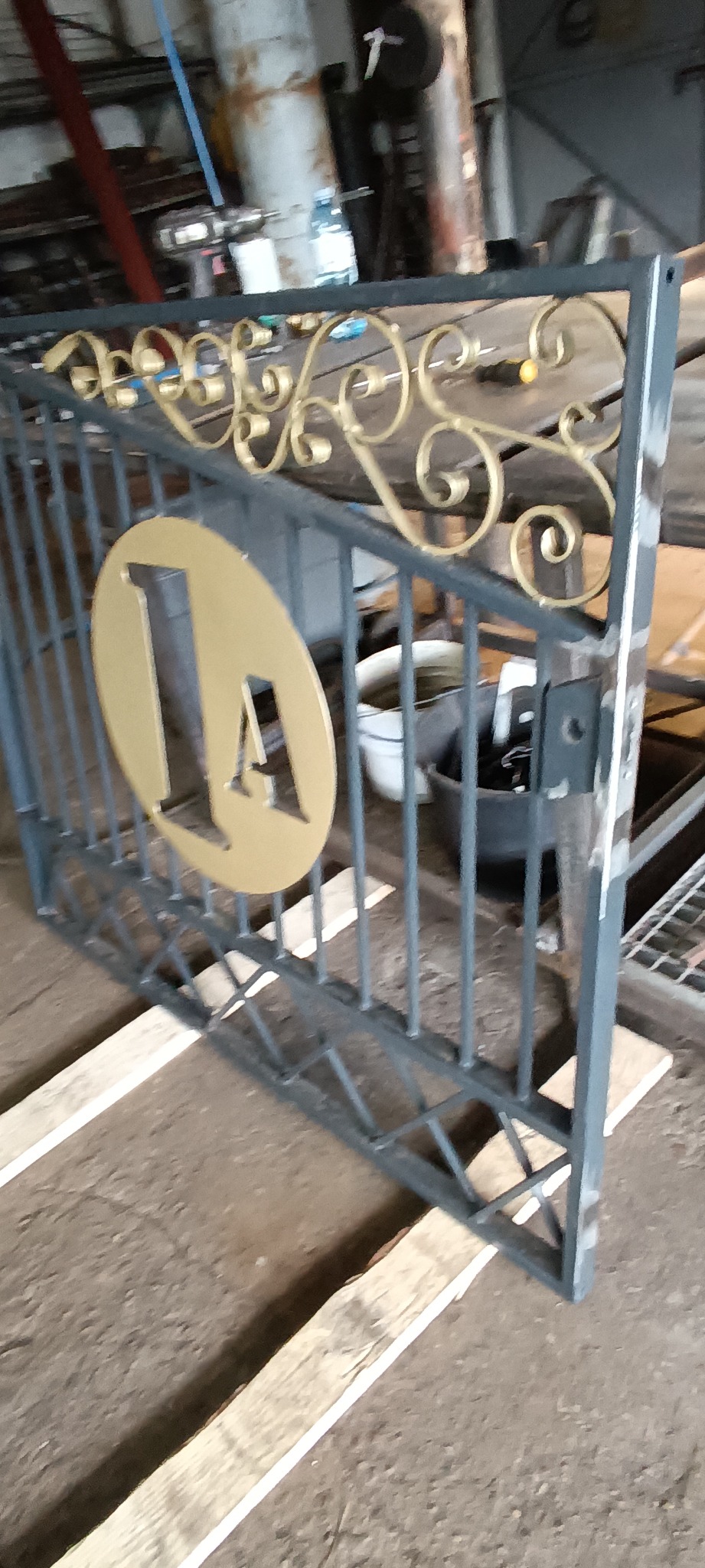 Fragment metalowej bramy w warsztacie, szara konstrukcja z dekoracyjnymi, złotymi elementami w kształcie spirali i owalu z literą 'A', wsparta na drewnianych belkach, w tle widoczne narzędzia...