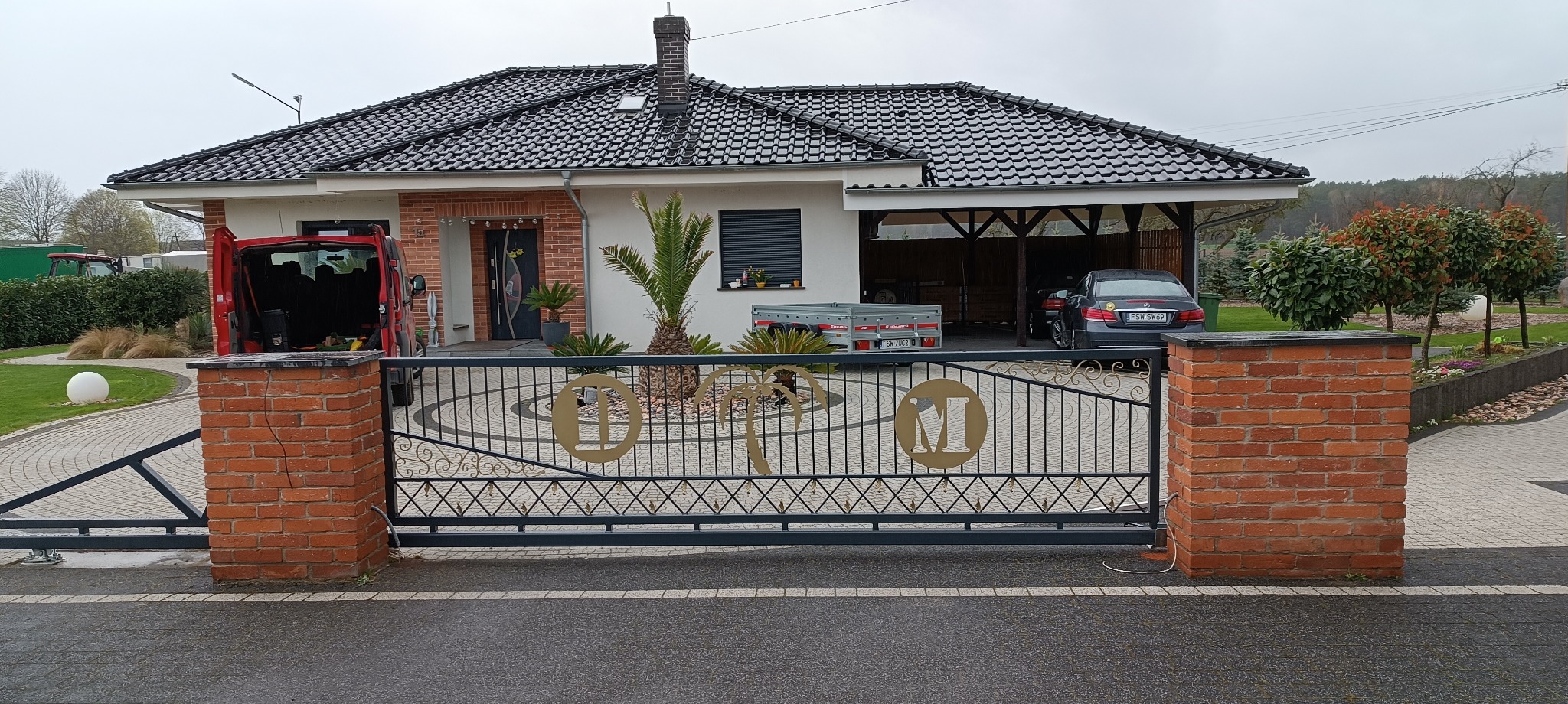 Automatyczna brama wjazdowa z ceglanymi słupkami i dekoracyjnymi elementami z motywem palm i liter, na tle domu z podjazdem i ogrodem.