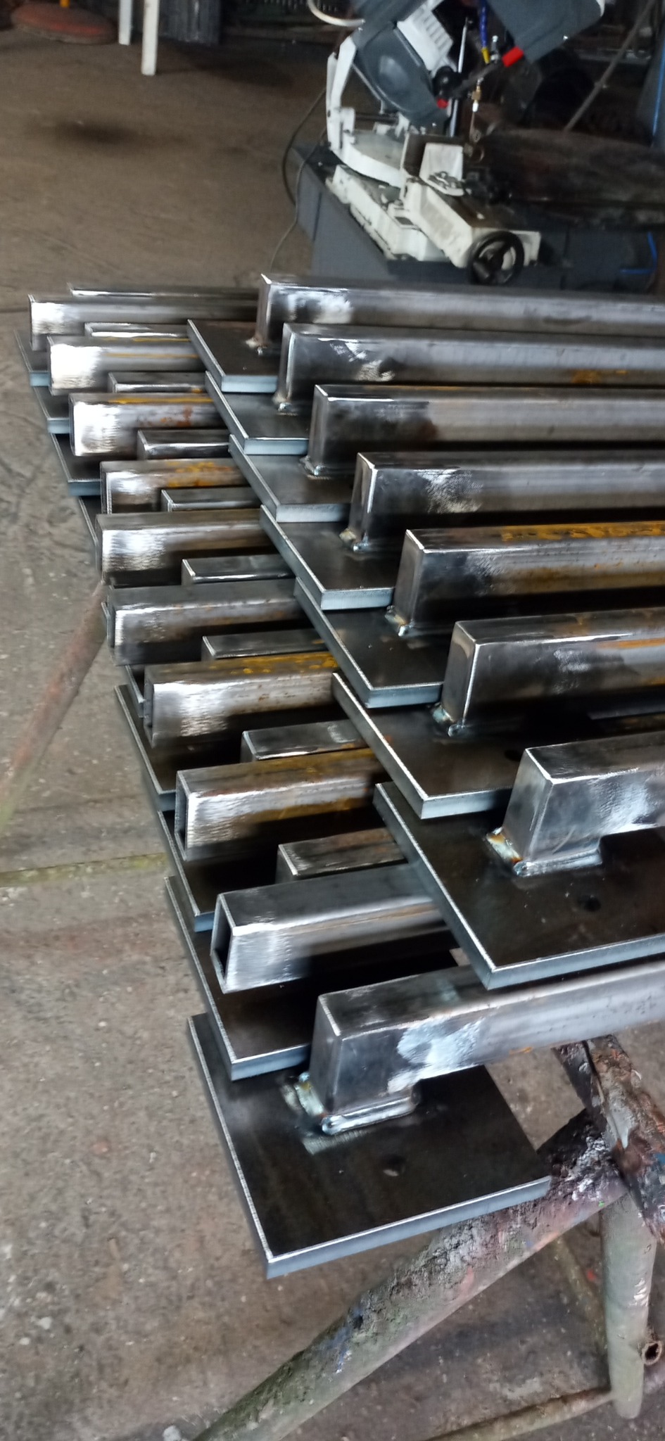 Stos metalowych elementów konstrukcyjnych, spawanych do kwadratowych podstaw, widocznych ślady spawania i obróbki metalu, w tle maszyna do cięcia metalu.