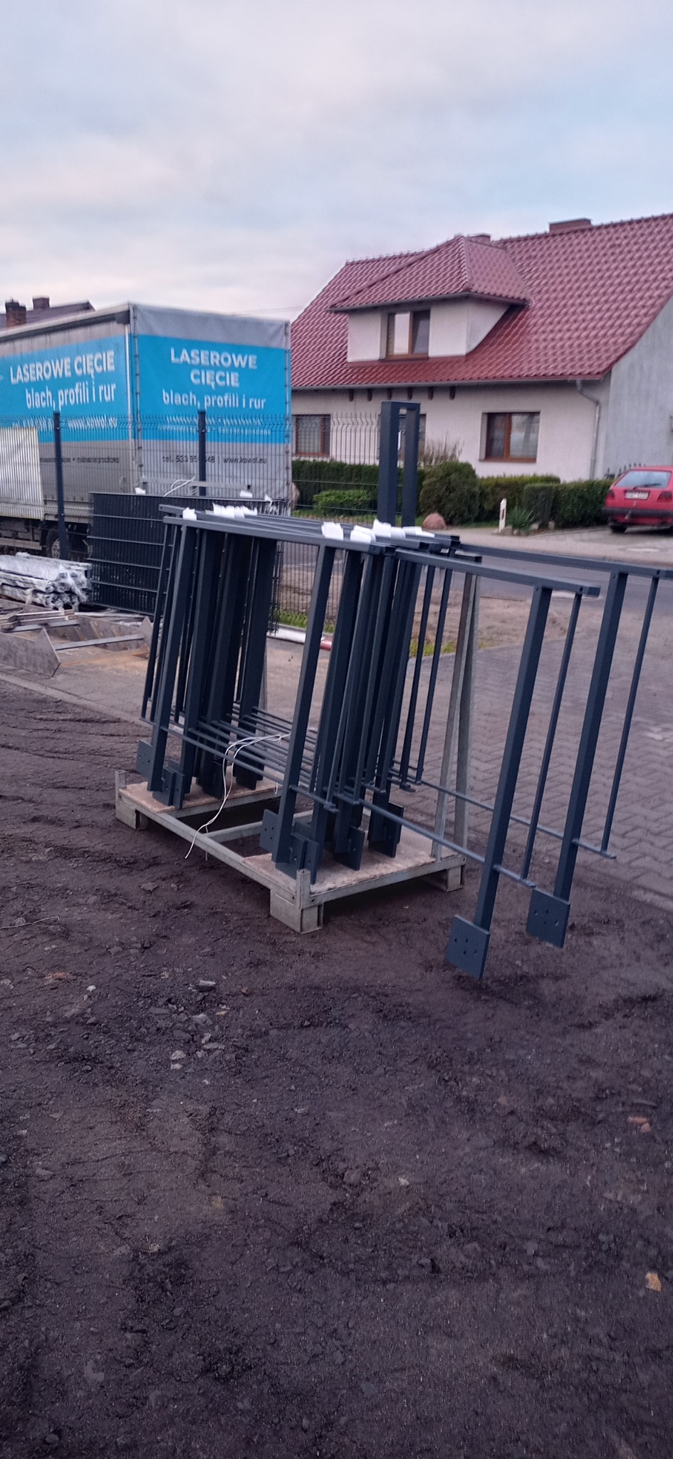 Ciemnoszare, metalowe elementy ogrodzenia, przygotowane do montażu, ustawione na palecie. W tle widoczny budynek mieszkalny z czerwonym dachem i samochód osobowy oraz ciężarówka z napisem 'Laserowe...