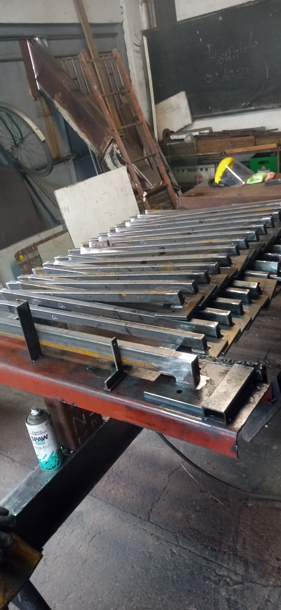 Stos metalowych profili przygotowanych do spawania, leżących na czerwonym stole warsztatowym, obok puszka ze środkiem antyodpryskowym, w tle widoczna drabina i tablica.