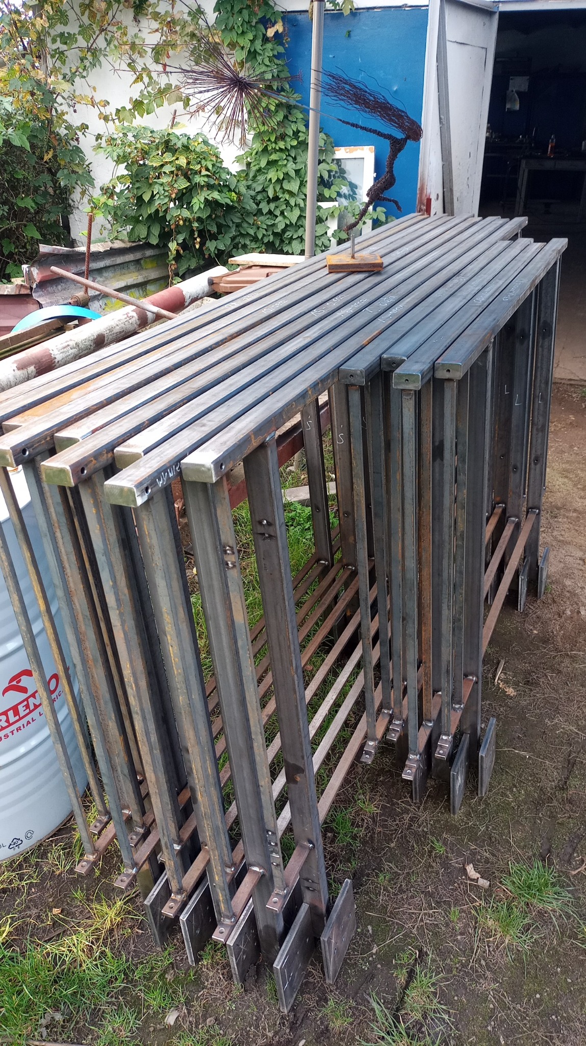 Stos metalowych, spawanych konstrukcji kratowych z kwadratowych profili, przygotowanych do montażu, z widocznymi śladami spawania i oznaczeniami, leżących na trawie przed warsztatem z dekoracyjną...