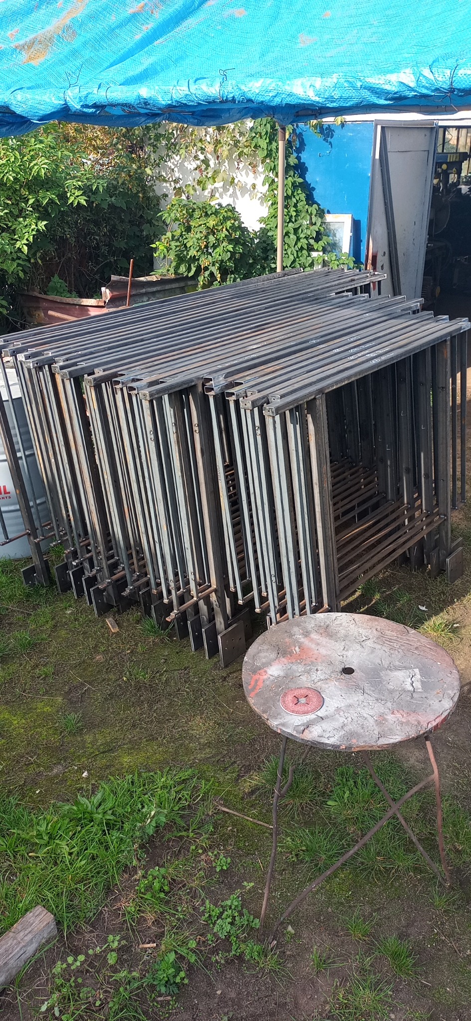 Stos surowych, spawanych, metalowych ram z profili kwadratowych z przyspawanymi płaskownikami, przygotowanych do dalszej obróbki lub montażu na zewnątrz warsztatu, obok okrągłego, metalowego...