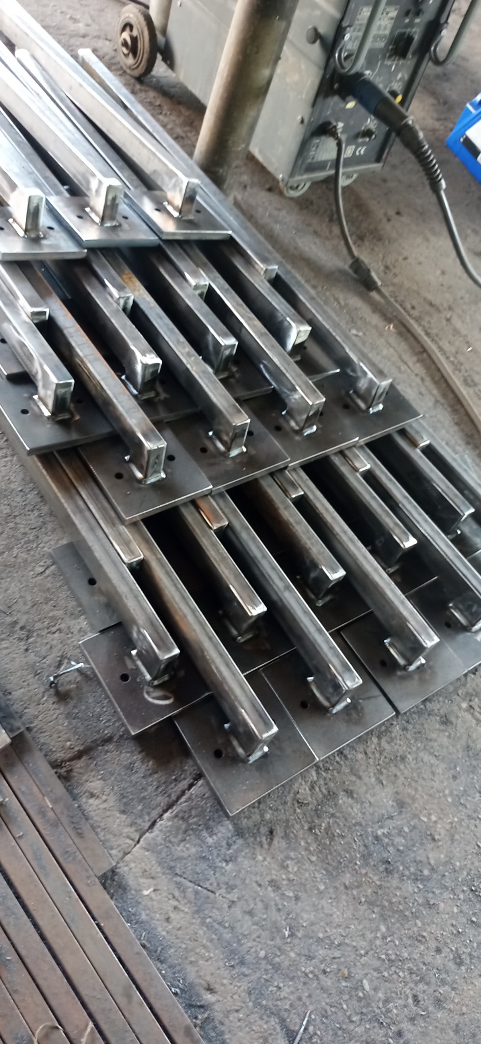 Przygotowane elementy konstrukcyjne ze stali: spawane profile kwadratowe do płaskowników z otworami, widoczny sprzęt spawalniczy w tle.