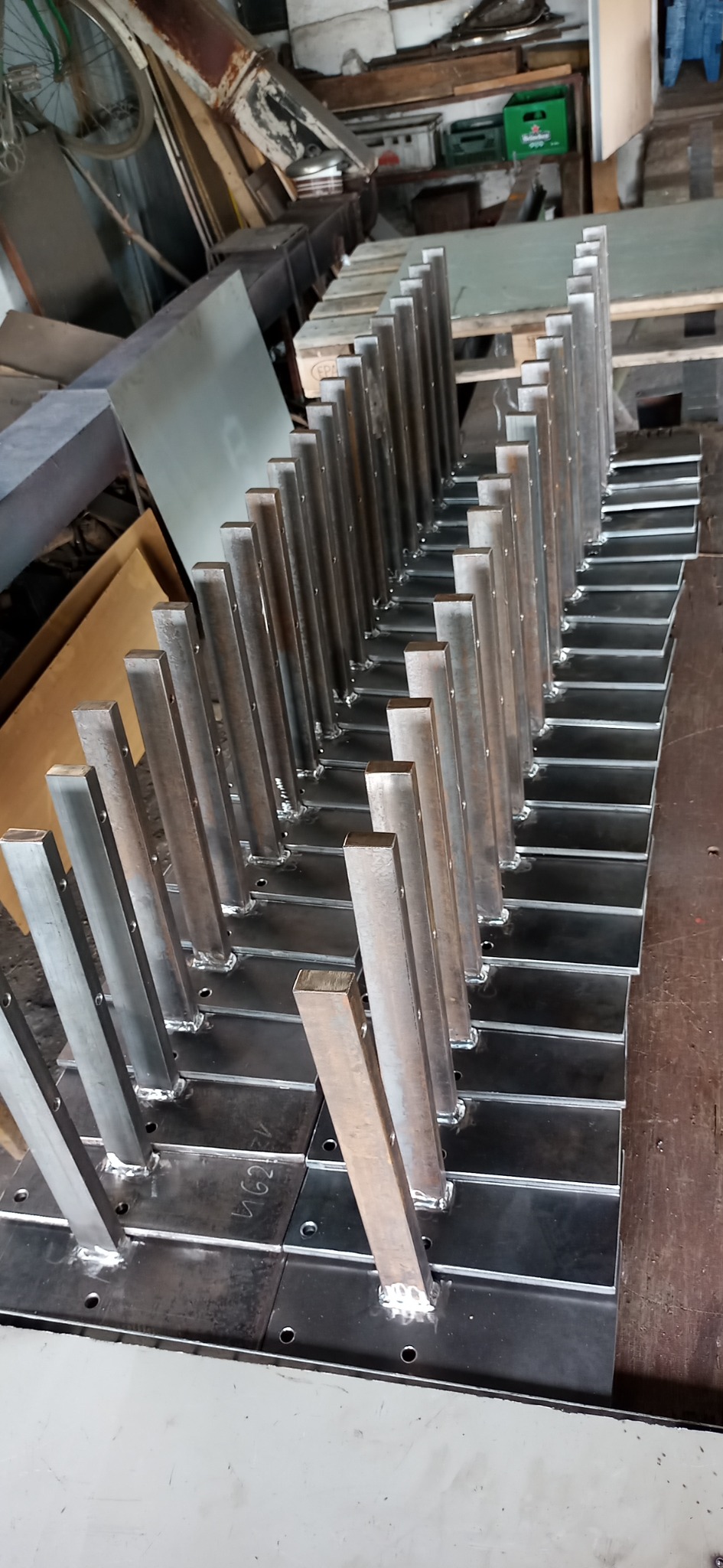 Rząd spawanych, stalowych słupków z kwadratowymi podstawami, przygotowanych do dalszej obróbki lub montażu, widoczne ślady spawania, w tle warsztatowy nieład.