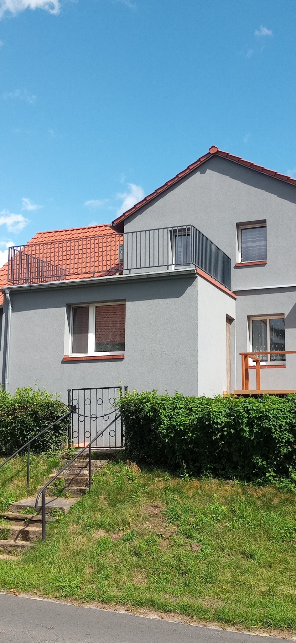 Szary dom jednorodzinny z metalową balustradą na dachu i balkonie, metalowa furtka wejściowa do piwnicy, betonowe schody, bujna zieleń, błękitne niebo.