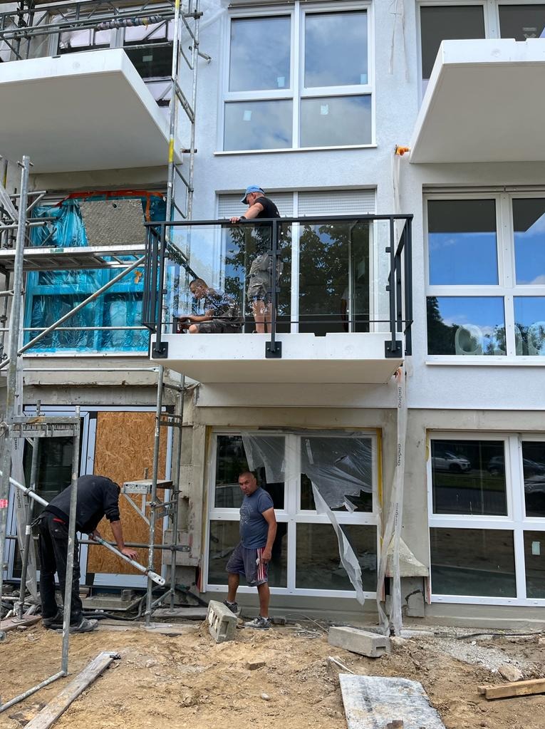 Montaż balustrady szklanej na balkonie nowego budynku, widoczni pracownicy na rusztowaniu i na balkonie, w tle okna z folią ochronną.