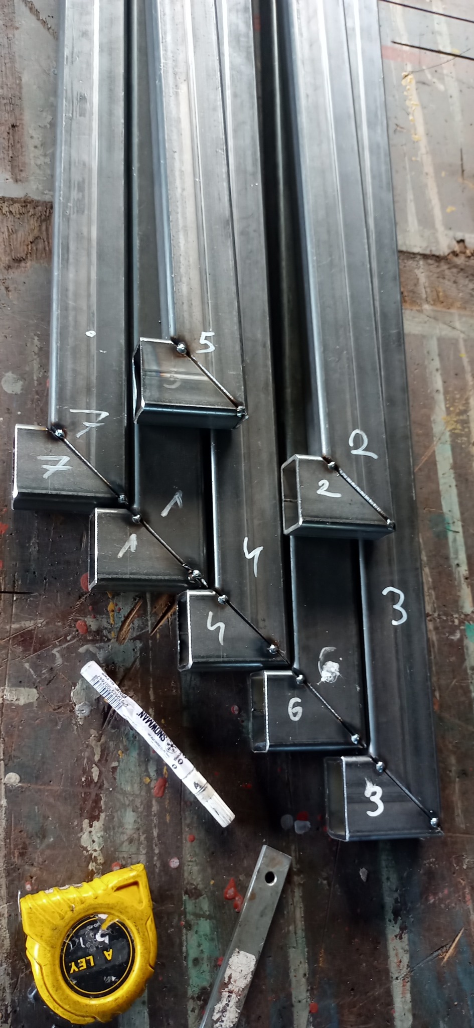 Przygotowane, ponumerowane profile metalowe z przyspawanymi elementami narożnymi, leżące na warsztatowym stole, obok miara zwijana i marker.