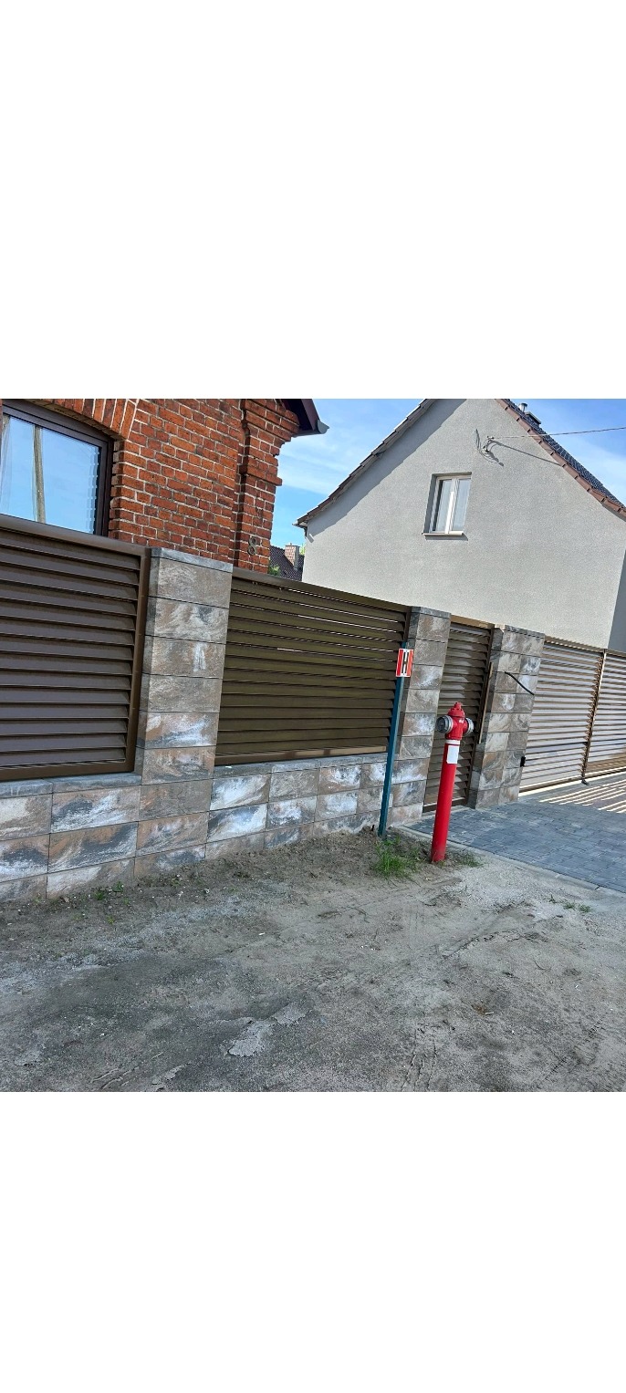 Nowoczesne ogrodzenie posesji z brązowymi, poziomymi panelami i kamienną podmurówką. Czerwony hydrant na pierwszym planie, budynki w tle.