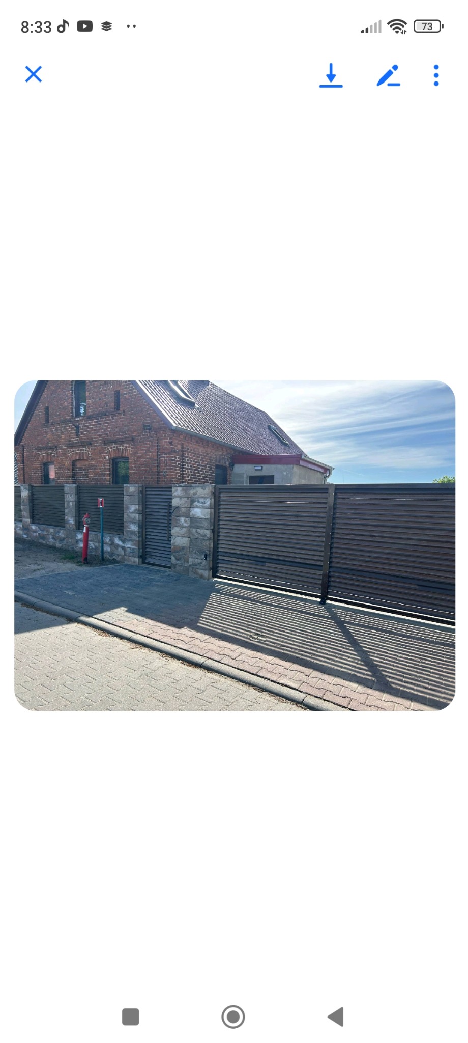 Nowoczesna brama wjazdowa z poziomych, brązowych paneli, osadzona na kamiennych słupkach. Widok na dom z czerwonej cegły i fragment chodnika.