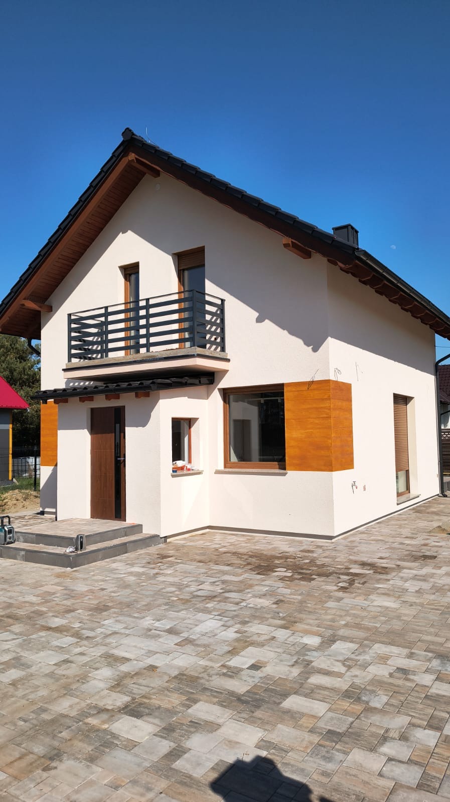 Nowoczesny dom z balkonem i czarną, metalową balustradą. Elewacja w odcieniach bieli i drewna, bruk na podjeździe. Błękitne niebo w tle.