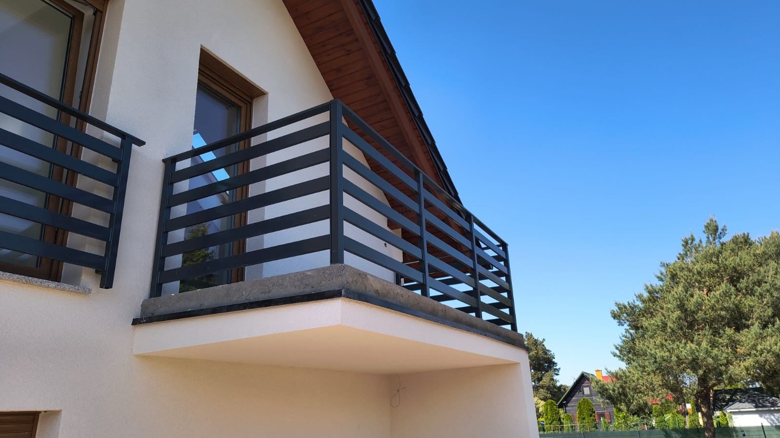 Nowoczesna, pozioma balustrada balkonowa w kolorze antracytowym, zamontowana na jasnym balkonie domu jednorodzinnego, widziana pod kątem z dołu, na tle błękitnego nieba.