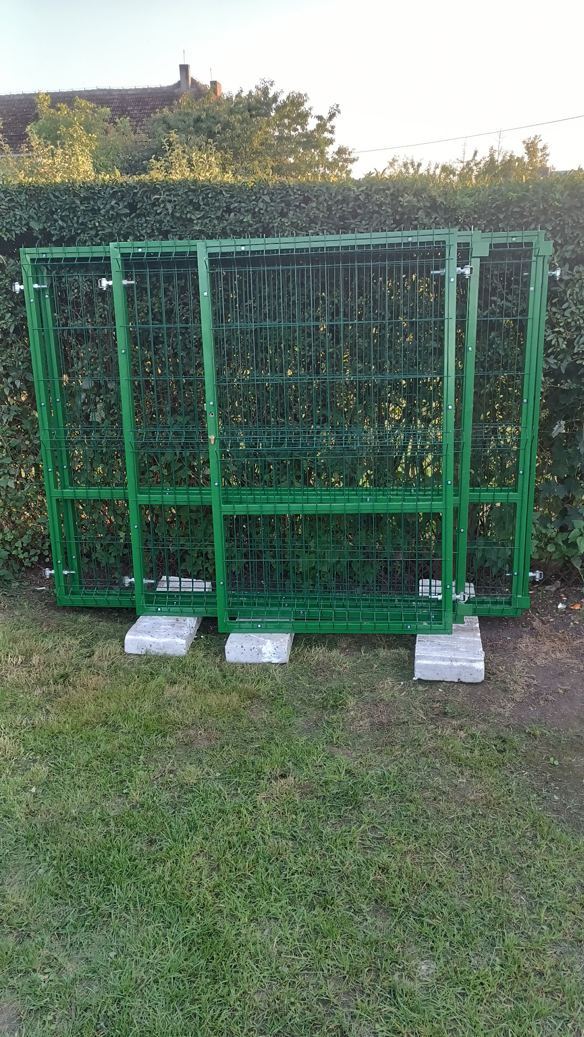 Zielone, metalowe przęsła ogrodzeniowe o siatkowej strukturze, oparte na betonowych bloczkach, na tle zielonego żywopłotu i trawnika.