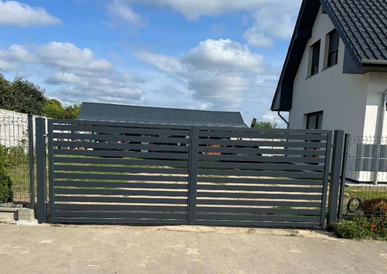 Nowoczesna, pozioma brama wjazdowa w kolorze grafitowym, z widocznym domem w tle i fragmentem ogrodzenia z siatki po lewej stronie.