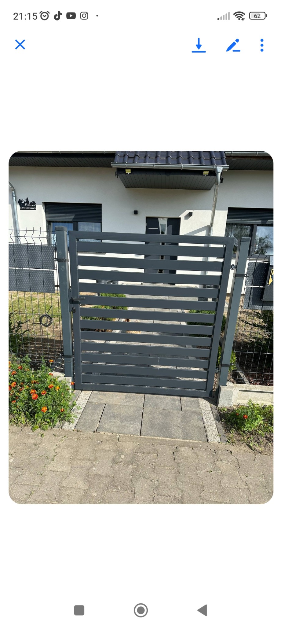 Szara, metalowa furtka z poziomymi szczeblami, osadzona w ogrodzeniu z siatki. Furtka prowadzi do domu z białej cegły, otoczona zielenią i kwiatami.