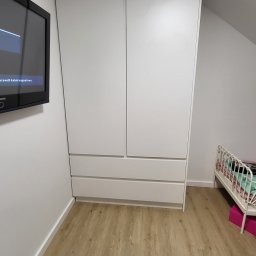 Stolarnia na wymiar - Biała szafa wnękowa z szufladami, dopasowana do skosu dachu, widoczna w pokoju obok łóżeczka dziecięcego i stolika z książką Harry Potter.