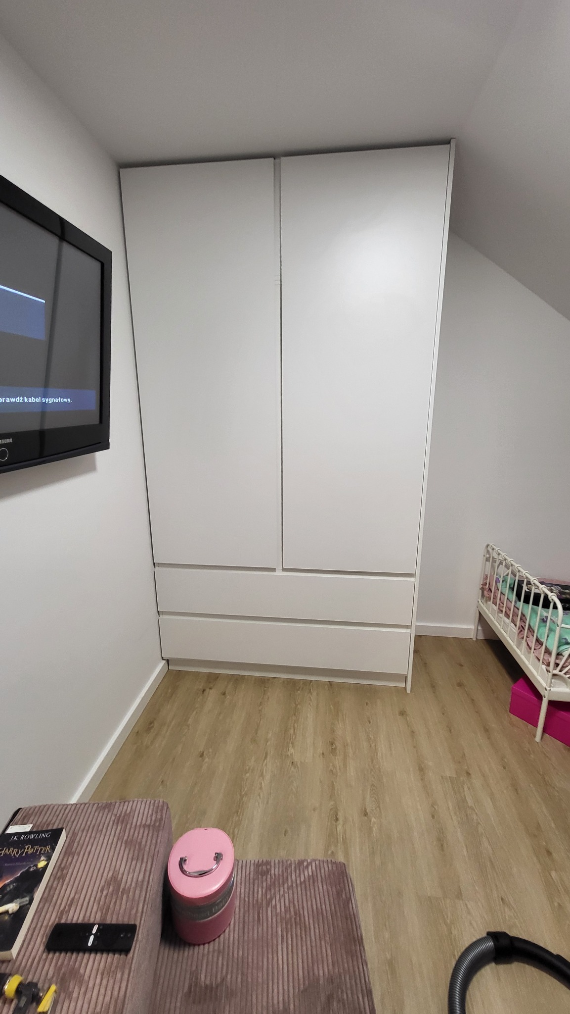 Biała szafa wnękowa z szufladami, dopasowana do skosu dachu, widoczna w pokoju obok łóżeczka dziecięcego i stolika z książką Harry Potter.