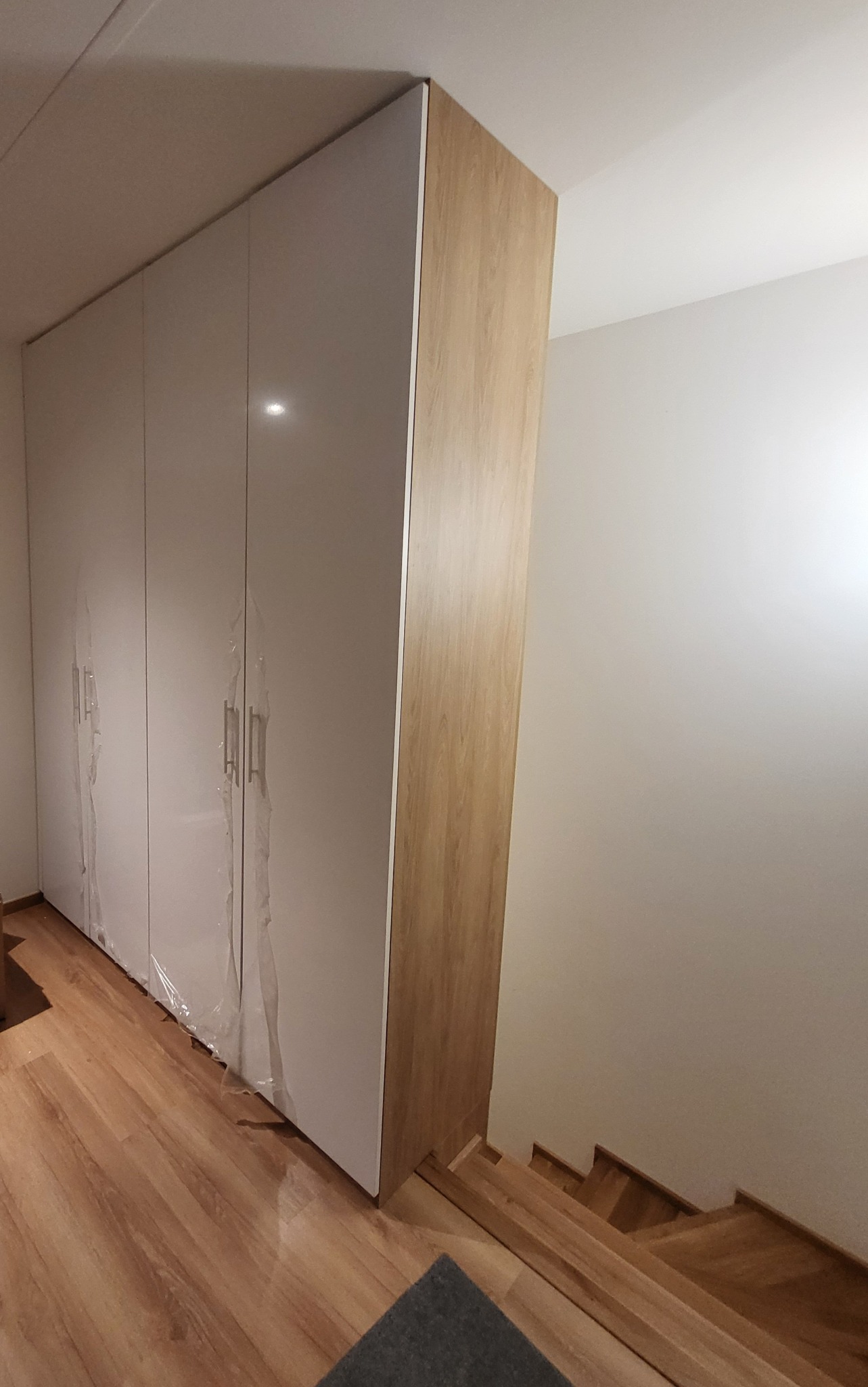 Nowoczesna szafa wnękowa z białymi frontami i drewnianym bokiem, częściowo zabezpieczona folią ochronną, widoczna z perspektywy podłogi z parkietem i fragmentem schodów.