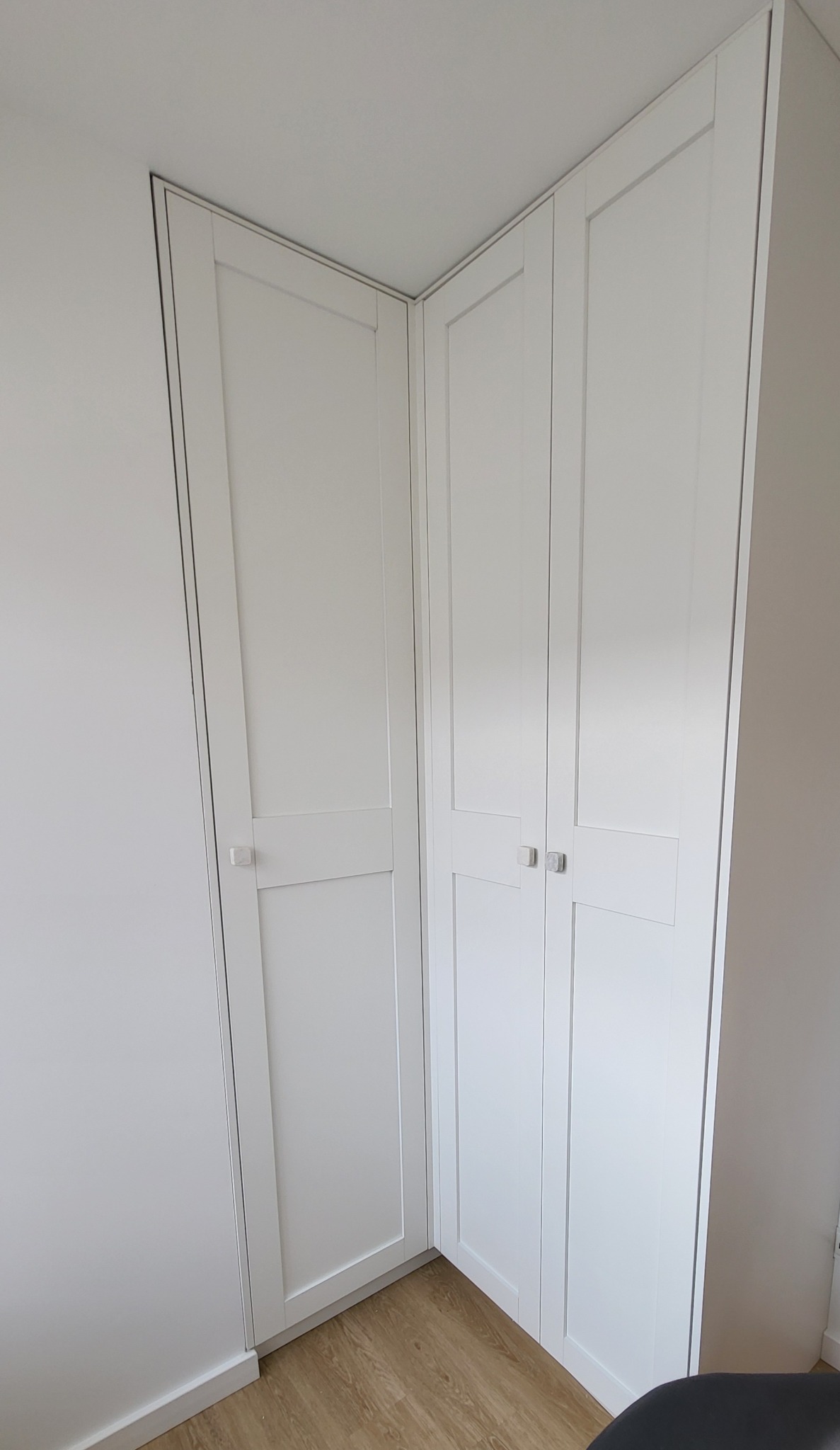 Biała szafa wnękowa narożna z dwiema parami drzwi, widoczne minimalistyczne uchwyty i fragment podłogi z paneli w odcieniach drewna.