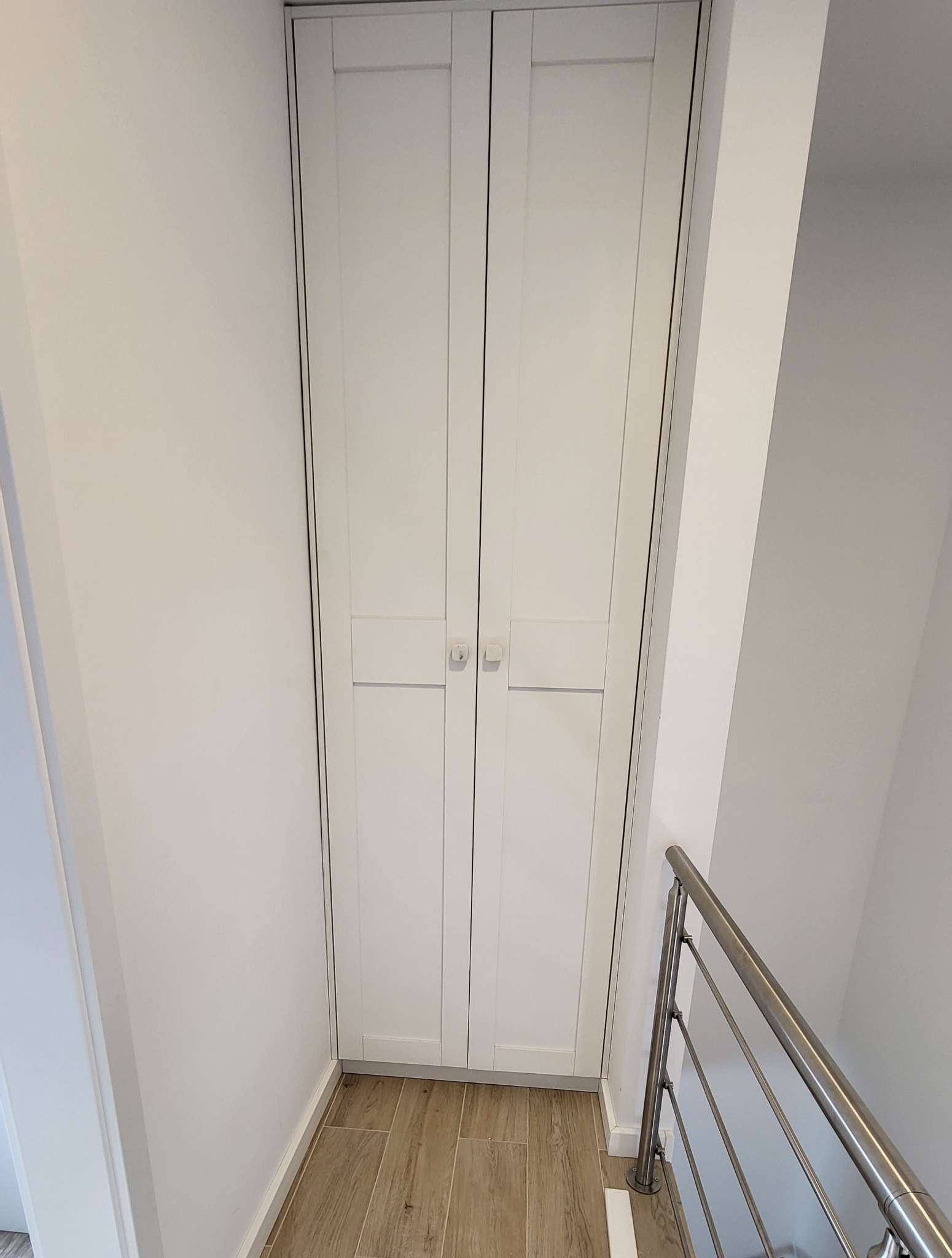 Biała, dwudrzwiowa szafa wnękowa z minimalistycznymi uchwytami, wkomponowana w przestrzeń pod schodami z drewnopodobną podłogą i metalową balustradą.
