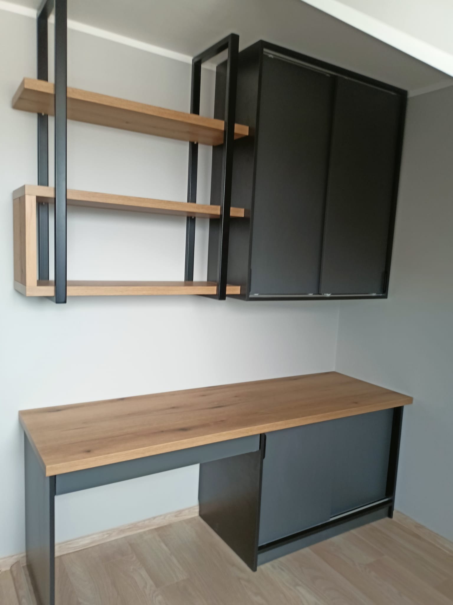 Nowoczesny zestaw mebli: biurko z drewnianym blatem i szarą podstawą, nad nim półki z drewnianymi blatami i czarną metalową ramą oraz szafka z przesuwnymi drzwiami.