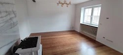 Puste wnętrze po remoncie w Niemczech, widoczna drewniana podłoga, białe ściany, okno z zasłonami i dekoracyjny grzejnik pod oknem, w rogu pomieszczenia kominek z marmurową obudową.
