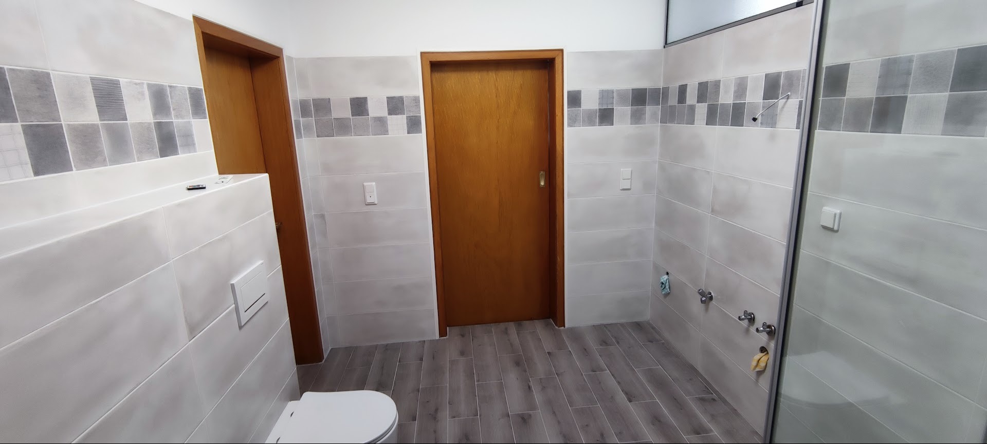 Wykończona łazienka z szarymi płytkami imitującymi drewno na podłodze, jasnoszare płytki ścienne z dekoracyjnym pasem szarych kwadratów, dwie drewniane ościeżnice drzwiowe i biała toaleta w rogu.