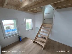 Wnętrze domu po remoncie w Niemczech: jasny pokój z drewnianą podłogą, schody prowadzące na antresolę, dwa okna z widokiem na zewnątrz, białe ściany i sufit z boazerii.