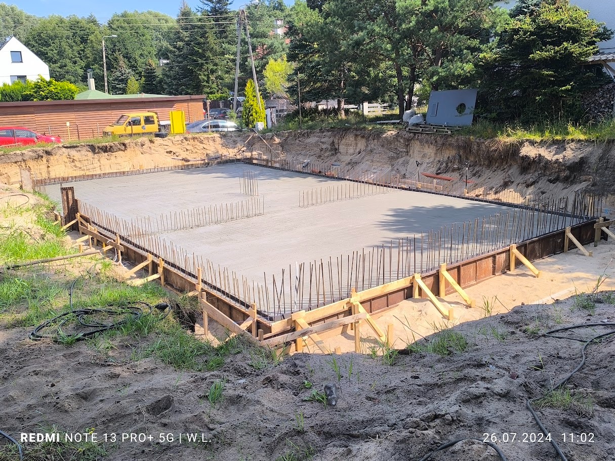 Wylany fundament pod budynek jednorodzinny, widoczne zbrojenie i szalunki, teren przygotowany pod budowę, w tle zabudowa jednorodzinna i drzewa.