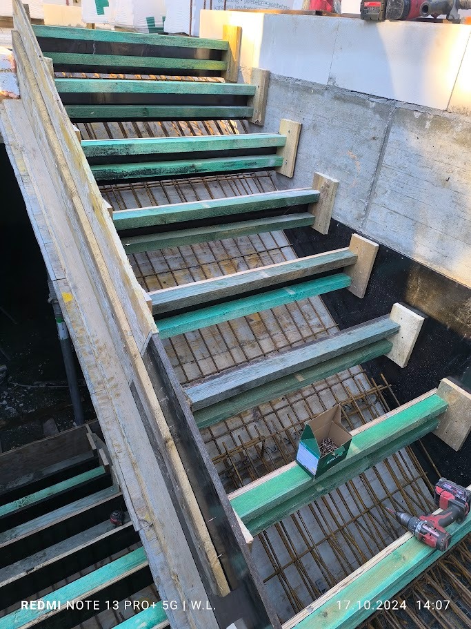 Szczegółowe ujęcie szalunku schodów z widocznym zbrojeniem, drewnianymi elementami konstrukcyjnymi, wiertarką akumulatorową i pudełkiem gwoździ na budowie domu.