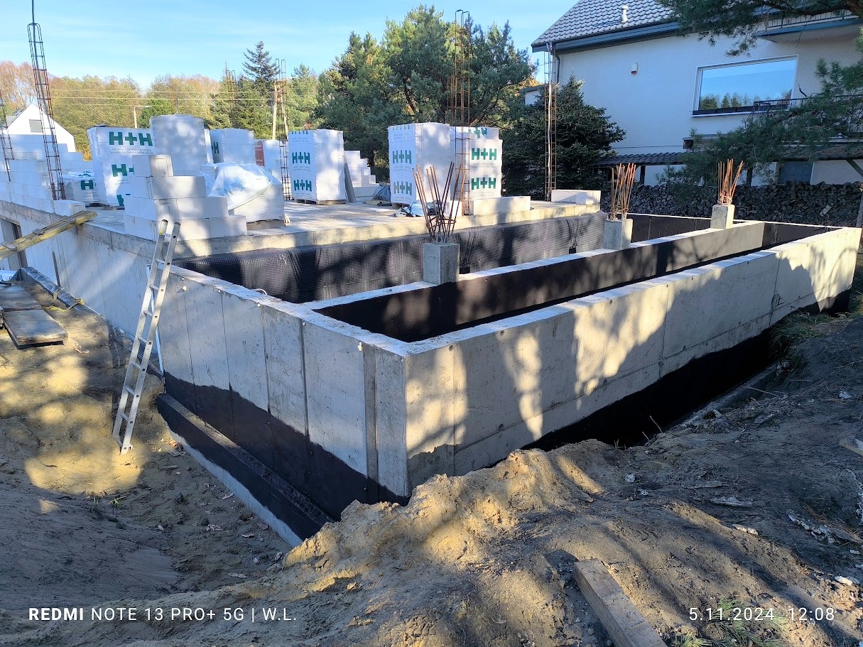 Fundamenty domu jednorodzinnego z izolacją pionową, widoczne bloczki H+H na płycie fundamentowej oraz wystające pręty zbrojeniowe, drabina oparta o ścianę fundamentową.