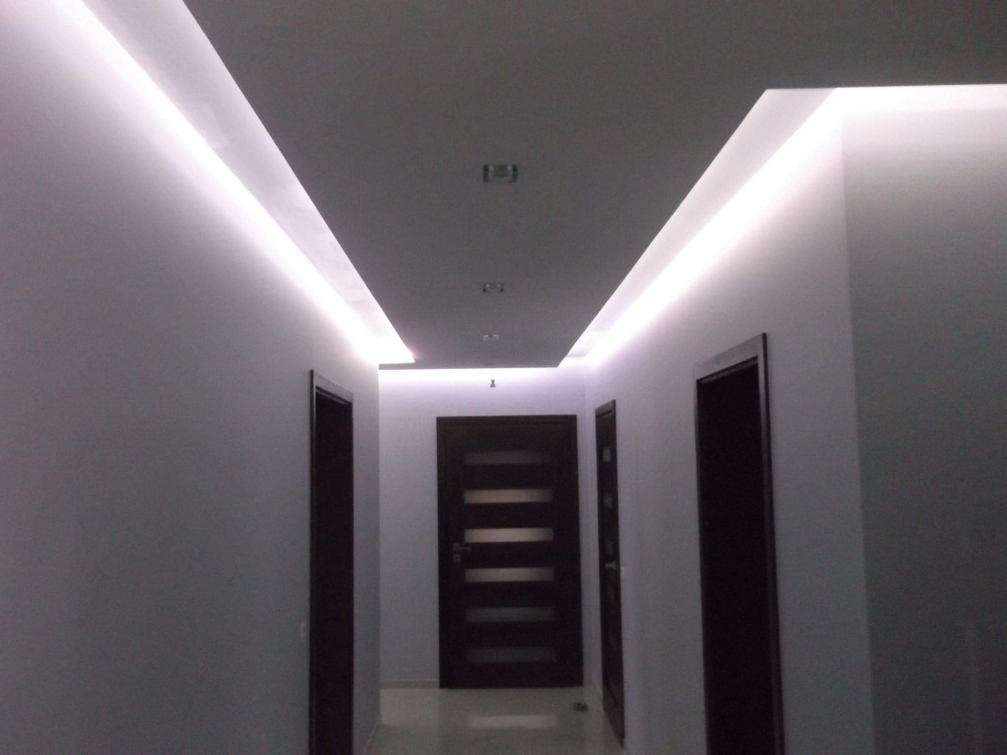 Korytarz z podwieszanym sufitem i oświetleniem LED, widoczne trzy ciemne drzwi z poziomymi przeszkleniami, szare ściany i podłoga z połyskiem.