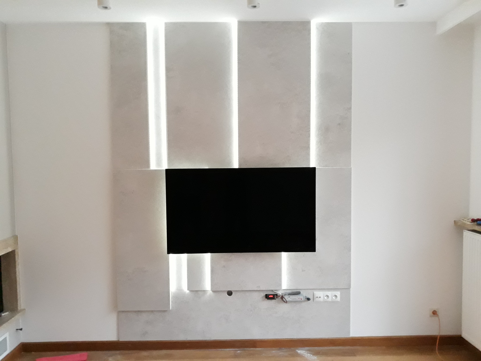 Ściana z dekoracyjnymi panelami oświetleniowymi i zamontowanym telewizorem, widoczne gniazdka elektryczne i listwa przypodłogowa.