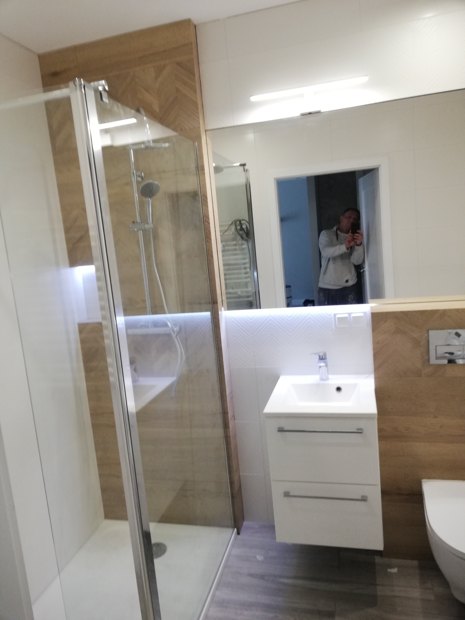 Nowoczesna łazienka z kabiną prysznicową typu walk-in, białą umywalką z szafką, toaletą i lustrem, w którym odbija się osoba robiąca zdjęcie.