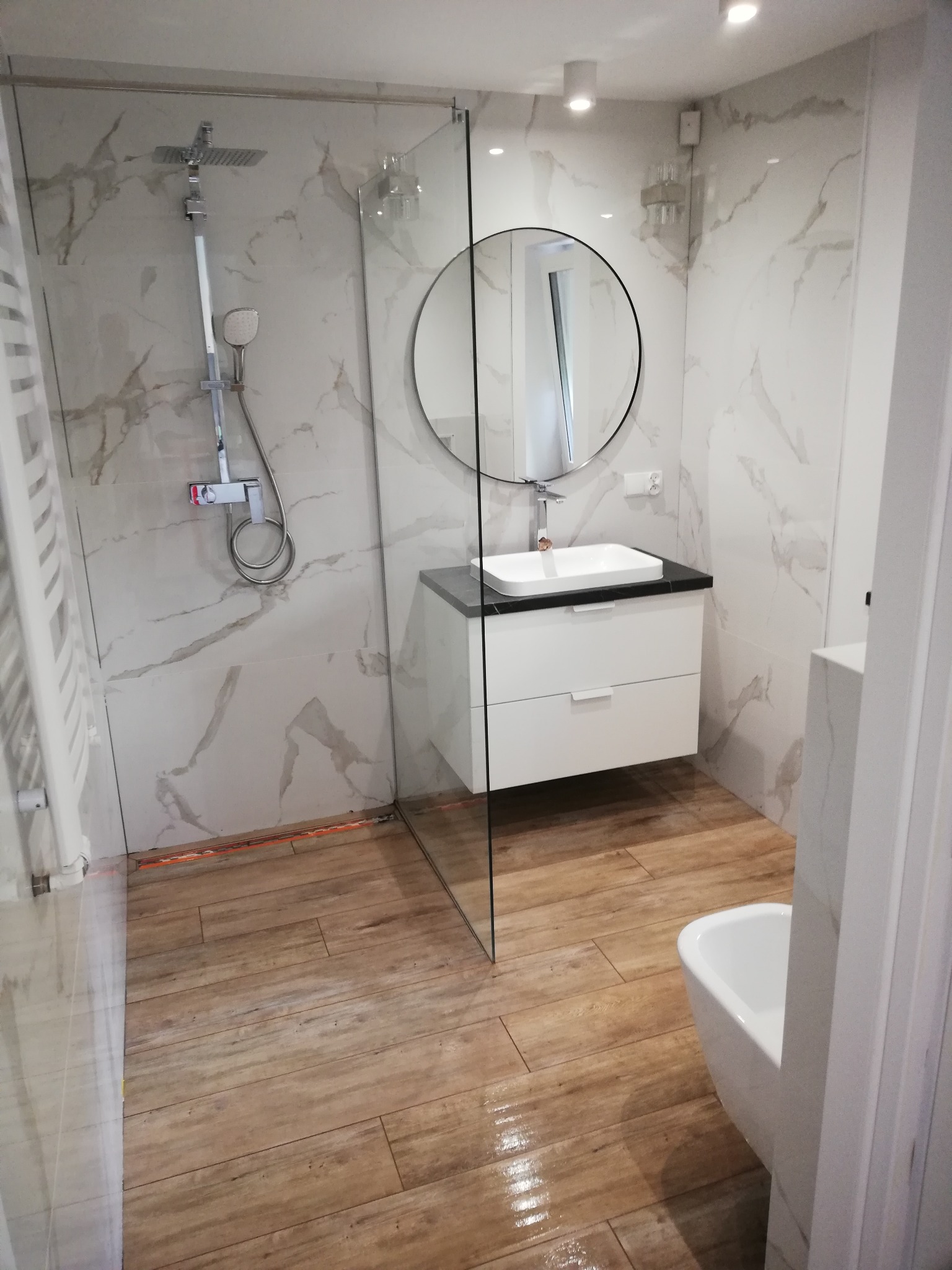 Nowoczesna łazienka z kabiną prysznicową typu walk-in, okrągłym lustrem nad białą szafką z umywalką, oraz płytkami imitującymi drewno na podłodze i marmur na ścianach.