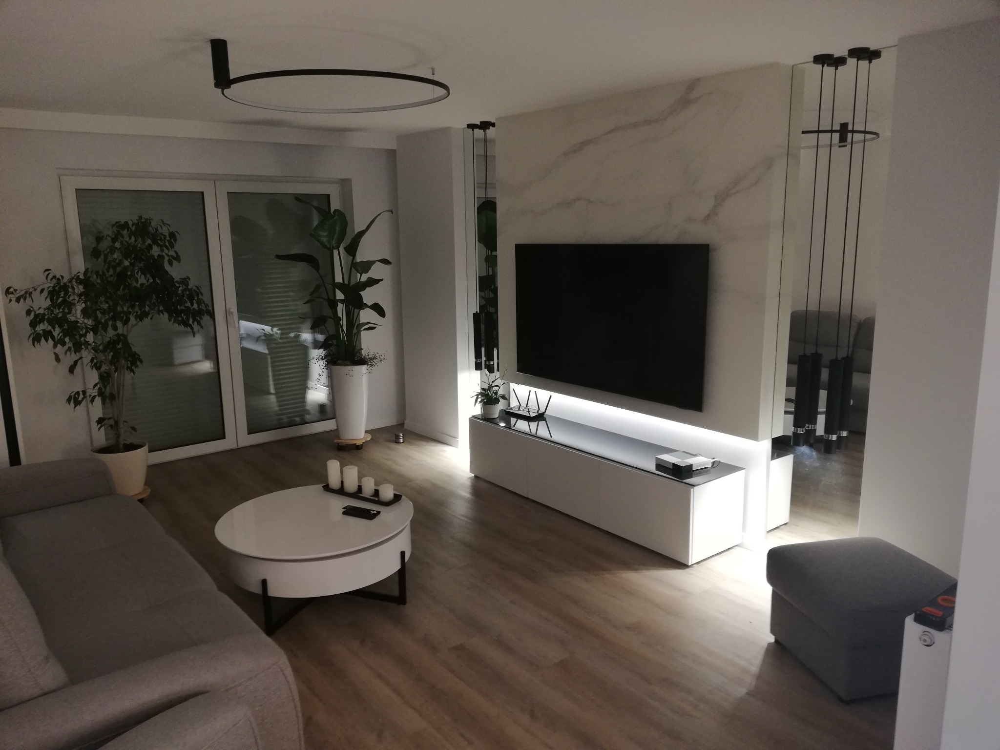 Nowoczesny salon z marmurową ścianą za telewizorem, podświetlaną szafką RTV, okrągłym stolikiem kawowym z dekoracyjnymi świecami i drewnianą podłogą.