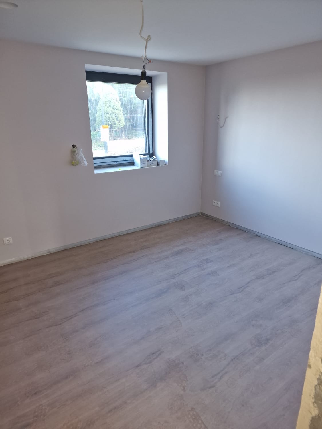 Wykończone wnętrze pokoju z panelami podłogowymi w odcieniach szarości, świeżo pomalowanymi ścianami na jasny kolor oraz oknem z widokiem na zewnątrz. Na ścianie widoczne ślady po taśmie malarskiej...