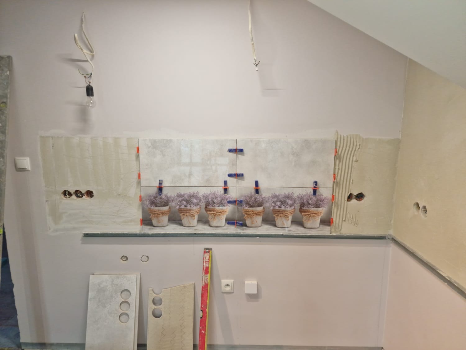 Ściana w trakcie układania płytek w łazience, z dekoracyjnymi doniczkami z lawendą na blacie, widoczne narzędzia i klej do płytek.