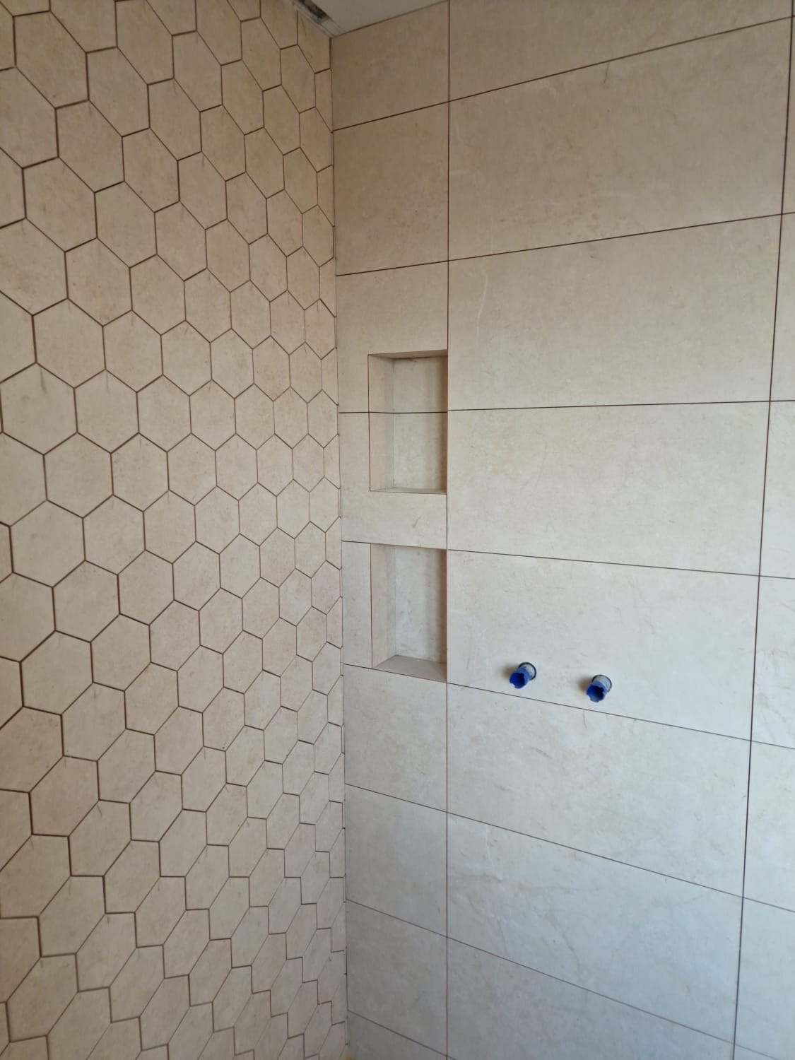 Fragment łazienki w trakcie wykańczania, z heksagonalnymi płytkami na jednej ścianie i dużymi, prostokątnymi płytami na drugiej, z wnękami w ścianie i wystającymi niebieskimi końcówkami rur.