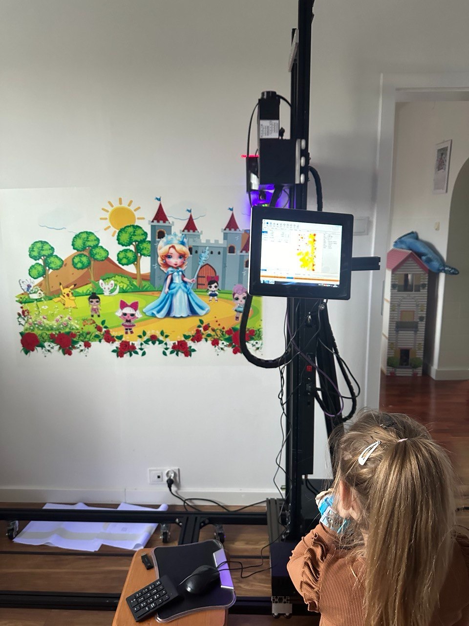 Dziecko obserwuje drukarkę ścienną, nanoszącą kolorową grafikę z motywem księżniczki na białą ścianę w pokoju.
