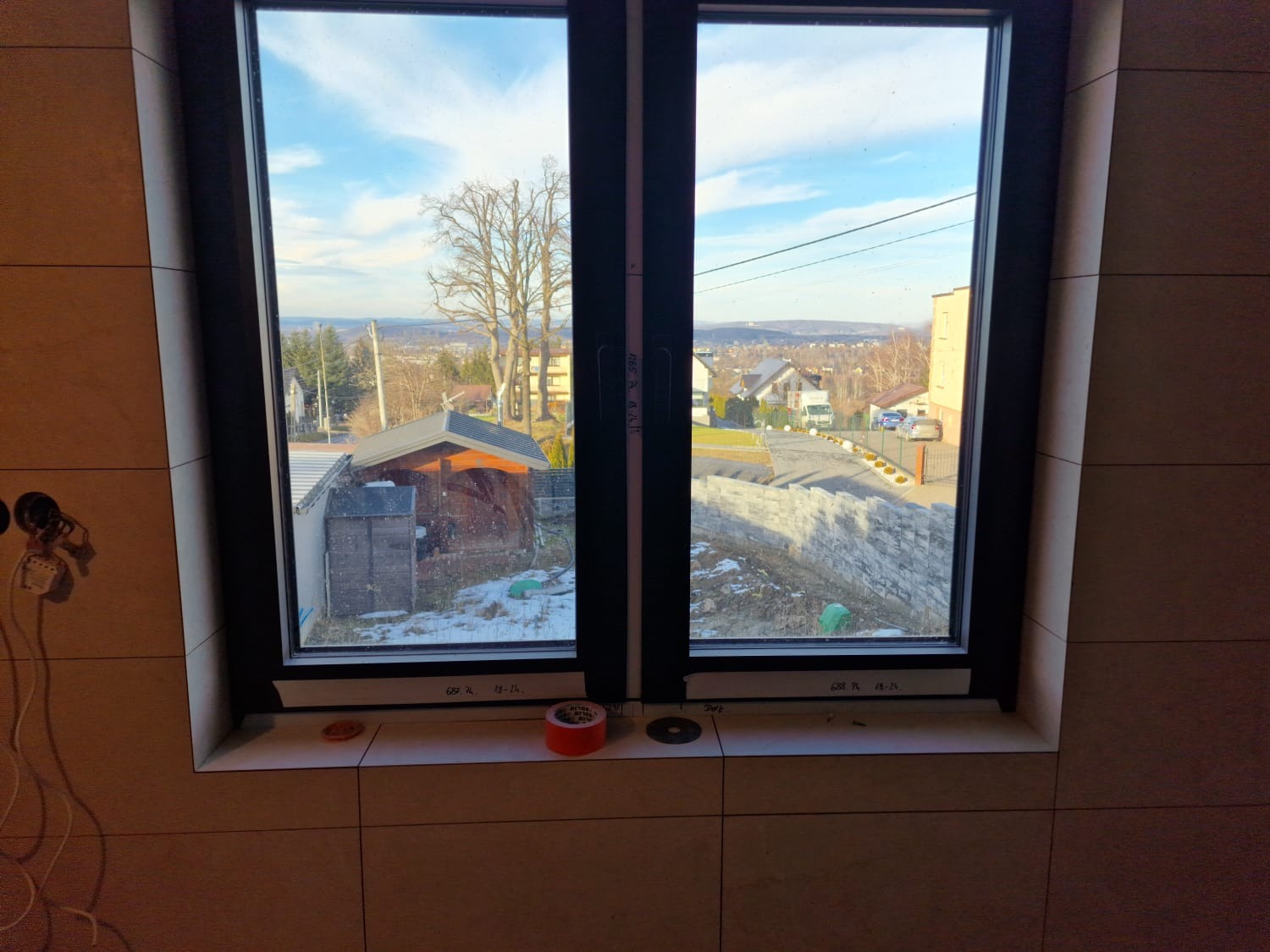 Zamontowane dwuskrzydłowe okno z czarną ramą, widok na zewnątrz z zabudową i częściowo ośnieżonym terenem, na parapecie resztki po montażu: taśma malarska, podkładki dystansowe, ściana wyłożona...