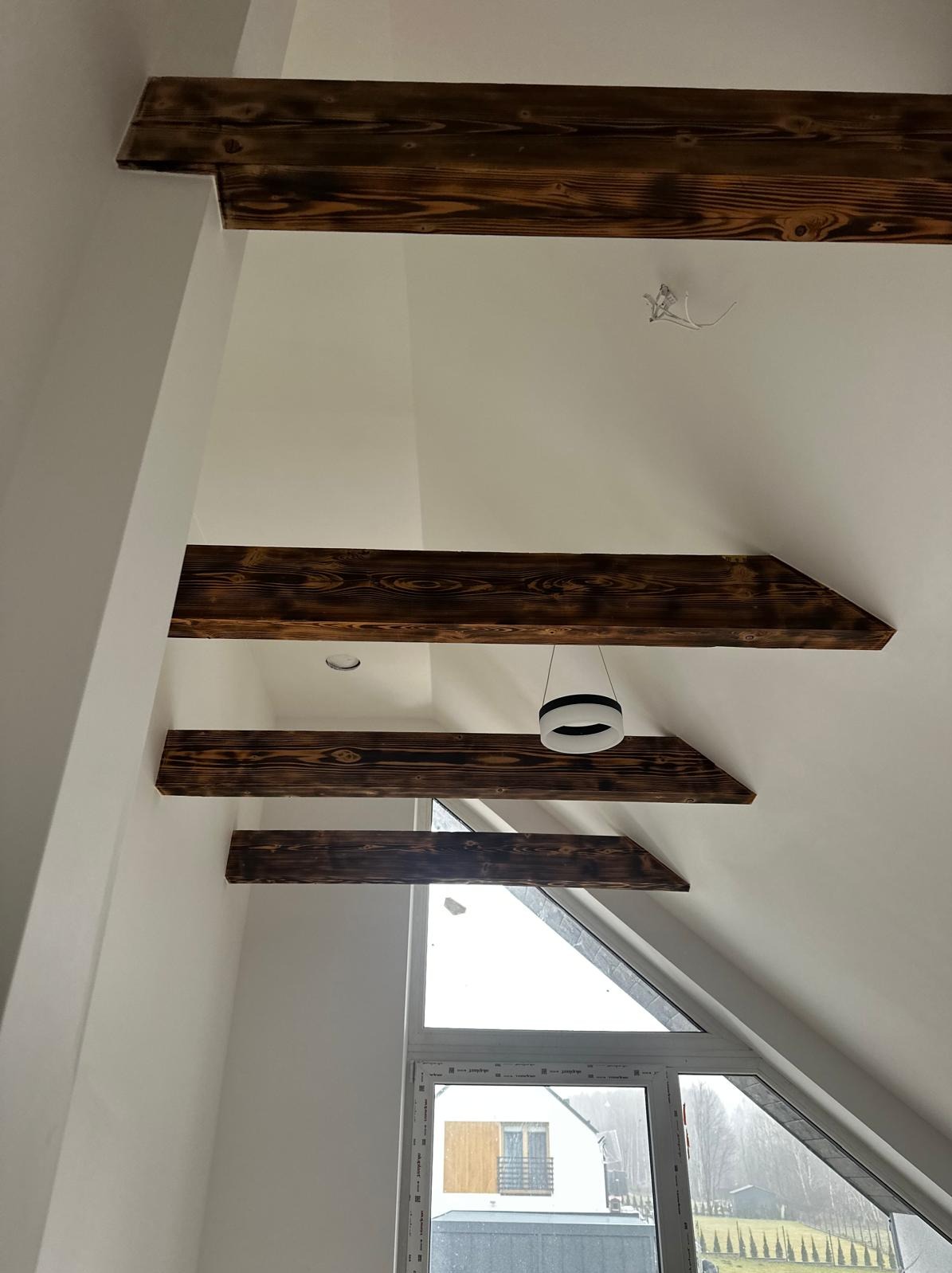 Skośne poddasze z drewnianymi belkami stropowymi o ciemnym, opalonym wykończeniu, widok na okno dachowe i nowoczesną lampę wiszącą.