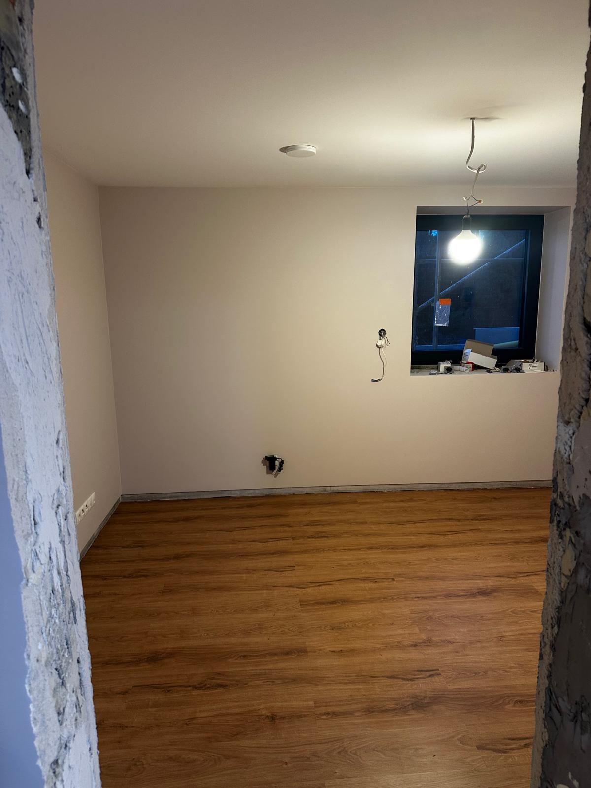 Pokój po malowaniu ścian na jasny odcień, z ułożonymi panelami podłogowymi o wzorze drewna, widoczne niedokończone gniazdka elektryczne i wisząca żarówka tymczasowa